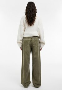 Femme aux longs cheveux bruns ondulés portant un pull crème et un pantalon en velours côtelé vert olive ample, debout de dos sur un fond blanc.