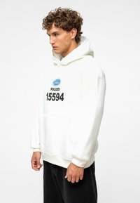 Witte hoodie met een voorzak, met zwarte tekst "POLIZEI 15594" en een blauw logo. Zacht materiaal en een losse pasvorm op een model.