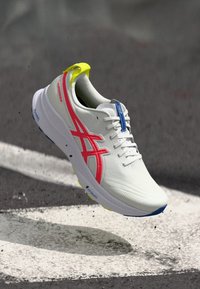 Running shoe in white with pink and blue accents. Light, textured material, round toe, and a bright yellow pull tab at the heel.