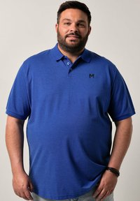 Modrá polo trička s krátkým rukávem vyrobená z hladké tkaniny, se třemi knoflíky na límci a černým logem na hrudi.