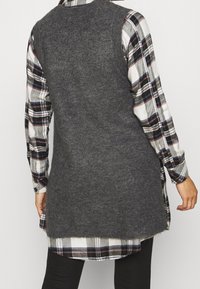 Grijze gebreide mouwloze vest over een zwart-wit geruit shirt, met een lange achterkant en een gestructureerde afwerking.