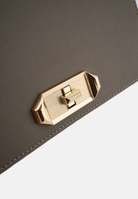 Ecke einer grauen, strukturierten Lederhandtasche mit einem vergoldeten rechteckigen Verschluss, der ein Rautenmuster und erhabene Akzente aufweist. Sichtbare Nahtdetails.