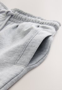 Gros plan sur un pantalon de survêtement gris montrant la taille élastique, le cordon de serrage et une poche latérale sur un fond neutre.
