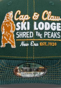 Gorra snapback de pana verde con un oso naranja bordado y texto blanco que dice "Cap & Claw Ski Lodge Shred the Peaks." Incluye el logo de New Era.