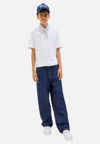 Un enfant se tient confiamment dans un t-shirt blanc, une écharpe bleue, un jean en denim et des baskets blanches, portant une casquette bleue sur un fond uni.