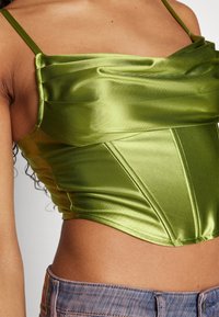 Top corto in satin verde con un design drappeggiato e texture, spalline sottili e dettagli delle cuciture strutturati. Indossato con jeans in denim chiaro.