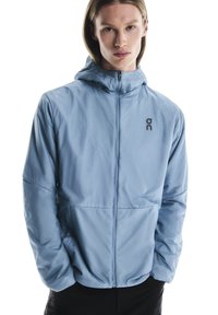 Veste légère bleu clair avec fermeture éclair intégrale, capuche réglable et accent logo sur la poitrine gauche. Texture lisse, design ajusté.
