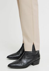 Pantalons de tailleur beiges avec fentes latérales à l'ourlet, associés à des bottines noires en cuir embossé crocodile avec bouts pointus et talons cubiques.