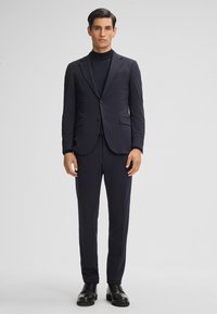 Abito blu navy con finitura testurizzata, vestibilità sartoriale; dolcevita e pantaloni coordinati; completato con scarpe nere stringate.