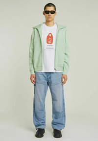 Haut à capuche vert menthe avec fermeture éclair, t-shirt blanc avec motif rouge, jeans large bleu clair, et baskets noires, portés par un mannequin.