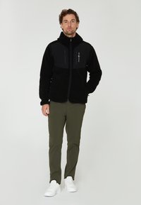 Zwarte fleecejack met een hoge kraag en ritszakken, gecombineerd met olijfgroene broek en witte sneakers op een neutrale achtergrond.