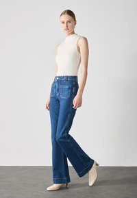 Jeans de campana azules de cintura alta, con dos bolsillos traseros, combinados con un body ajustado de canalé color crema y mules de tacón beige.