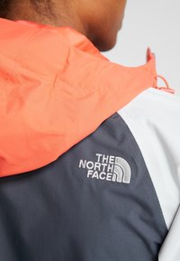 Personne portant une veste bicolore avec une capuche orange et un dos gris, avec le logo "The North Face" brodé dans le haut du dos.