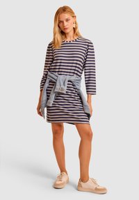 Pasiasta sukienka z długimi rękawami, w granatowe i srebrne paski, luźny krój, dżinsowa kurtka związana w pasie, w połączeniu z jasnymi sneakersami.