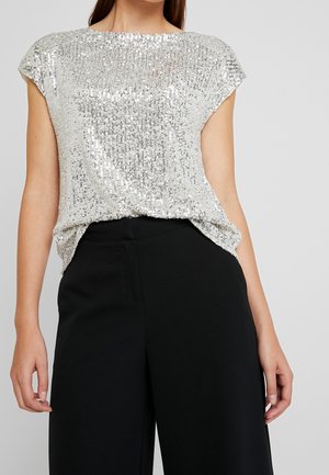 Femme portant un haut à manches courtes en sequins argentés, rentré dans un pantalon noir taille haute à jambes larges, sur un fond uni.