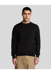 Felpa nera con collo a giro, maniche lunghe e polsini a costine. Presenta un logo sottile sul petto. Abbinata a pantaloni beige chiaro.