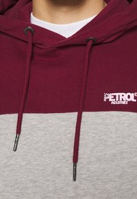 Sudadera bicolor burdeos y gris con capucha ajustable, textura suave y un logo en el pecho. Los cordones tienen puntas de metal.