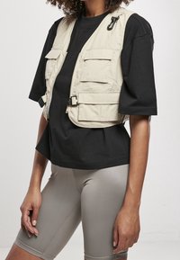 Beżowa kamizelka z wieloma kieszeniami, zamek błyskawiczny z przodu i klamrami, noszona na czarnej koszulce i szarych szortach rowerowych. Gładka faktura materiału.