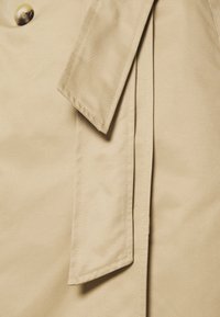 Blond trenčkot v detailu ukazující knoflík, límec a prošité poutko na opasek s detailním šitím.