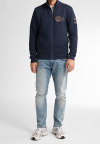 Marinblå zip-up jacka med texturerat tyg, med en rund logodetalj på bröstet och ribbade muddar och nederkant. Nedtonad tillsammans med ljusblå jeans.