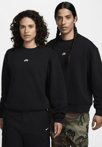 To personer har matchende sorte sweatshirts lavet af bomuld. Hver har et lille hvidt Nike-logo i midten. Baggrunden er ensfarvet hvid.