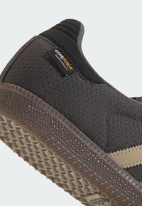 Zapatilla negra con una parte superior texturizada, suela de goma, detalles en marrón claro y una etiqueta de logo prominente cerca del talón.