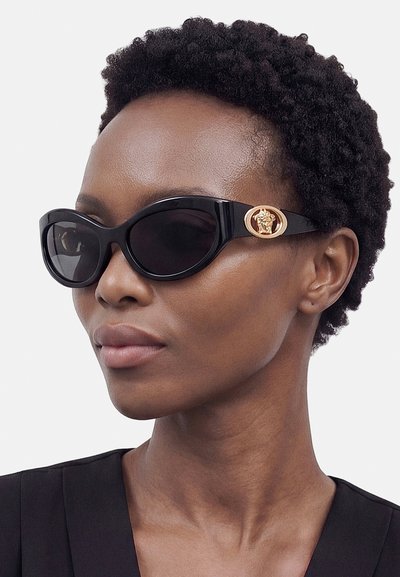 Donna con capelli corti e ricci che indossa occhiali da sole neri a forma di occhio di gatto con emblema dorato sul lato, vestita con un top nero.
