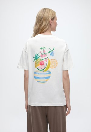 Γυναίκα που φοράει άσπρο t-shirt με πολύχρωμη απεικόνιση μπολ φρούτων στην πλάτη, που περιλαμβάνει ανανά, μπανάνα, φράουλες και εσπεριδοειδή.