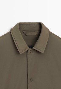 Veste vert olive avec un col classique, une fermeture à pression et une texture lisse. Présente un design minimaliste sans motifs ni accents visibles.