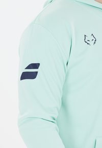 Mintgrøn hoodie med en glat tekstur, et rævlogo på brystet og en mørkeblå accent på venstre ærme.