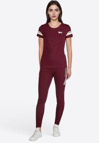 Camiseta atlética burdeos con rayas blancas, que presenta un logo de león. Combinada con leggins a juego que muestran "Londres" en fuente blanca.