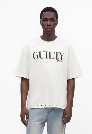 Giovane uomo che indossa una maglietta bianca oversize con la parola "GUILTY" in nero e un piccolo testo rosso sotto, abbinata a jeans azzurri.