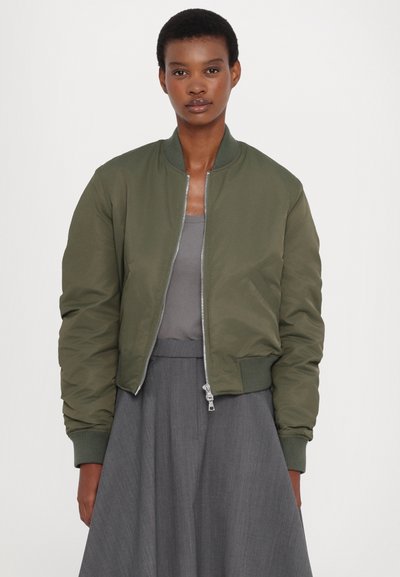 Olive green bomber jacket in a smooth material, zip-front closure, ribbed cuffs, and hem, paired with a grey tank and grey skirt.