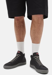 Beine mit schwarzen Shorts, weißen Sportsocken und schwarzen High-Top-Sneakern mit grauen Sohlen und Schnürsenkeln, stehend auf weißem Hintergrund.