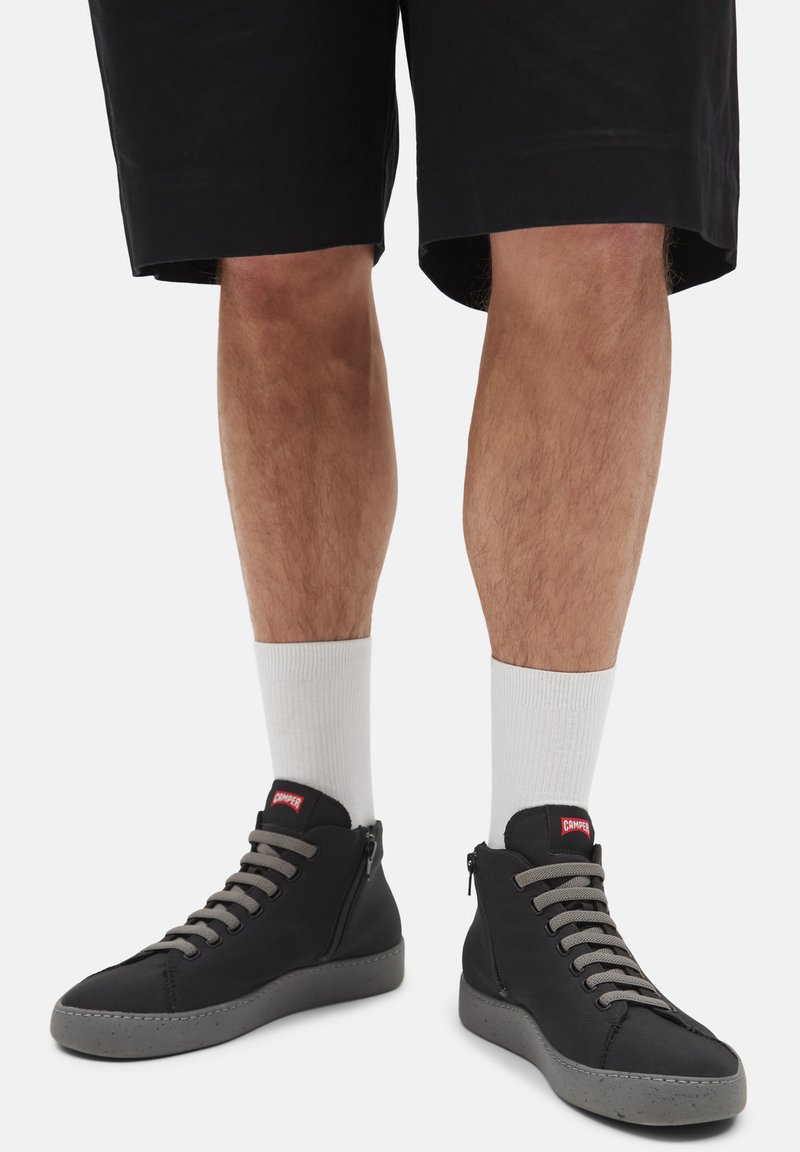 Sneakers negre cu glezna înaltă, cu șireturi gri, talpă texturată și logo roșu "Camper", asortați cu șosete albe și pantaloni scurți negri.