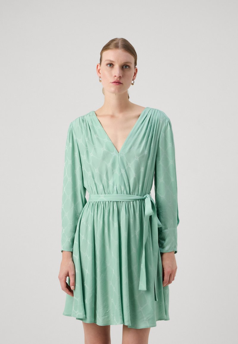 Rochie verde deschis cu mâneci lungi, decolteu în V, talie strânsă și curea legată. Prezintă un model subtil în diamante și un material moale, fluid.