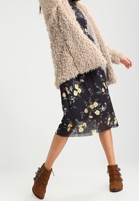 Femme portant une veste en fausse fourrure beige, une jupe midi noire à fleurs et des bottines à boucle marron, se tenant les jambes écartées sur un arrière-plan neutre.