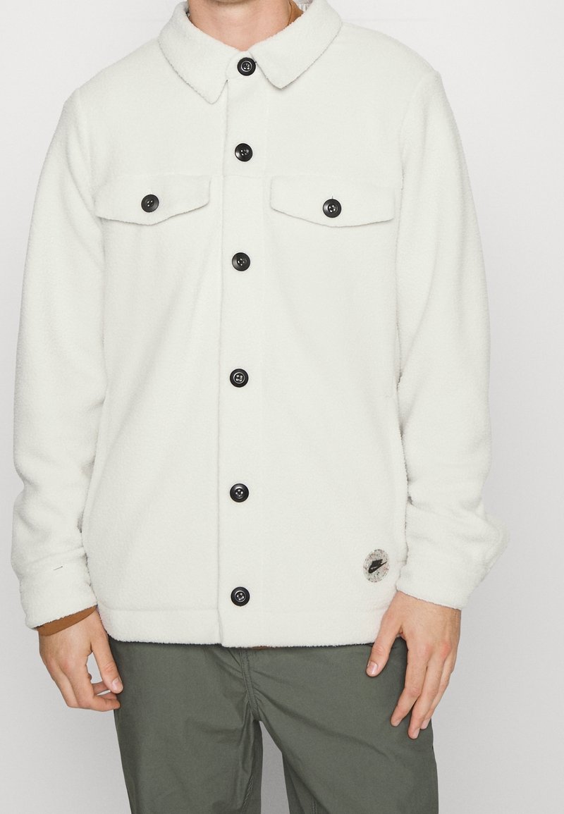 Vit fleecejacka med knappar och två bröstfickor, bärs med olivgröna byxor och en brun skjorta under.