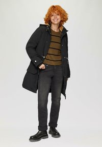 Schwarze Parka mit Kapuze, ausgestattet mit silbernen Beschlägen und Taschen. Darunter ein braun gestreifter Pullover, kombiniert mit schwarzen Jeans und Chelsea-Stiefeln.