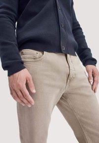 Beige katoenen jeans met een rechte snit, gecombineerd met een marineblauwe cardigan met een knopenfront en geribbelde manchetten. Hand geplaatste op de heup.
