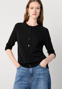 Svart knappopp cardigan med rund halsutringning, lange ermer og en myk strikket tekstur. Kombinert med lyseblå denimjeans.