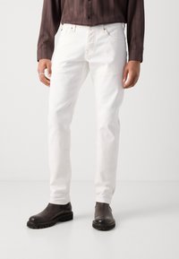 Jean en denim blanc avec une coupe slim, présentant un design classique à cinq poches et des accessoires en métal, associés à des bottines noires.
