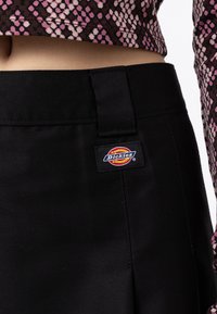 Čierne nohavice Dickies s hladkým povrchom a pútkom na pás, s farebnou nášivkou loga na boku. Látka vyzerá trvácne.