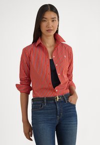 Červenobielo pruhovaná košeľa na zapínanie s rozšíreným golierom, vyhrnutými manžetami a malým detailom loga, v kombinácii s tmavomodrými džínsami.