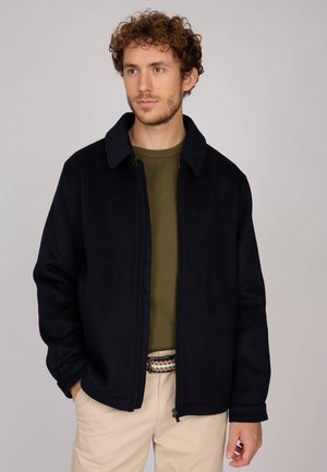Veste polaire zippée bleu marine avec col, portée sur un t-shirt en tricot olive, associée à un pantalon beige et une ceinture à motifs.