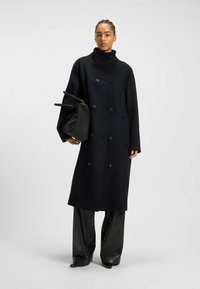 Donna che indossa un cappotto nero doppiopetto, pantaloni neri a gamba larga, mentre tiene una borsa nera su uno sfondo grigio uniforme.