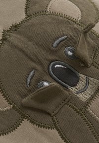 Khaki-Stoff mit einem Teddy-Bären-Gesicht-Design und gestickten Details, darunter graue Augen und braune Flecken, weiche Textur und Ohren.