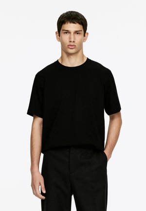 Schwarzes kurzärmliges T-Shirt aus Baumwolle; verfügt über einen Rundhalsausschnitt und eine lockere Passform. Kombiniert mit schwarzen Hosen, minimalistischem Design.