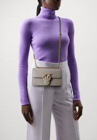 Borsa grigia con dettagli in oro, caratterizzata da un design unico della chiusura, abbinata a un dolcevita viola e pantaloni bianchi a vita alta.