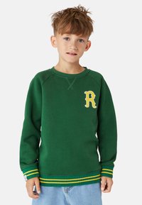 Grön sweatshirt med gula detaljer, ribbade ärmslut och nederkant, med en broderad gul bokstav "R" på bröstet. Mjuk material, ledig passform.
